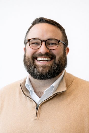 A smiling photograph of Patrick in front of a white background. He is wearing a yellow sweater and glasses. He has a beard and short brown hair.