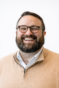 A smiling photograph of Patrick in front of a white background. He is wearing a yellow sweater and glasses. He has a beard and short brown hair.