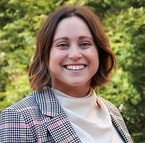 A smiling photograph of Kayla Poole in front of some trees. She has short brown hair, a pale pink top, and a grey and pink blazer on.